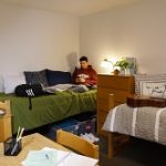 student sitting on bed in dorm room looking at phone