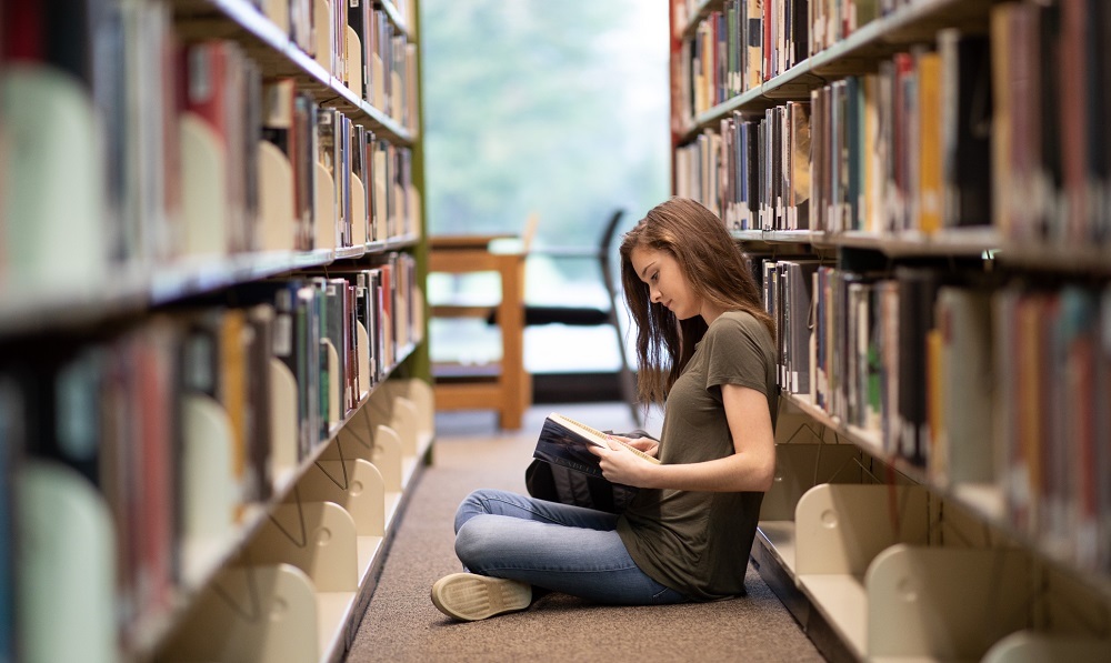 student sitting on floor in library reading a book