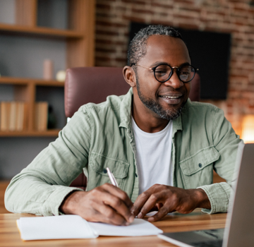 man sitting at laptop and writing in notebook